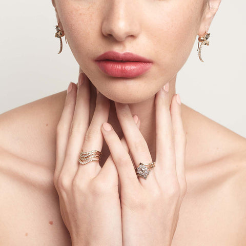 A cluster engagement ring with a rose cut champagne diamond center stone and round white diamond stones surrounding it set in yellow gold stacked with nesting bands on a models left hand and on her right there are white diamond curved bands. 3