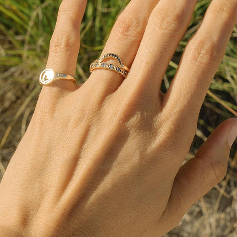 A 3mm straight band with white diamonds set in eternity around the band set in yellow gold stacked beneath a crescent shaped band on a light-complextion hand. On her pinky finger is an "A" monogrammed signet ring. 4