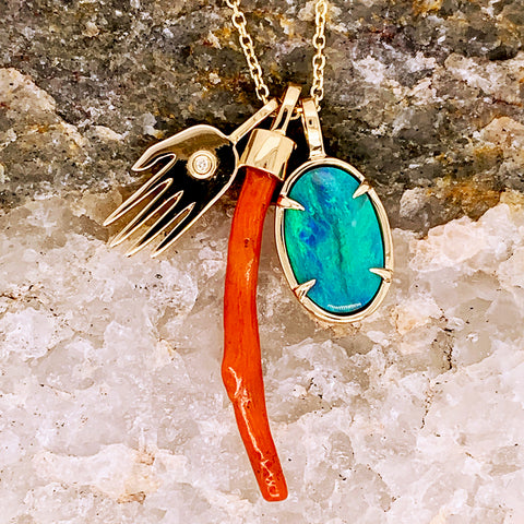 An oval one of a kind turquoise charm with a coral charm and a hand charm with a white diamond accent in yellow gold on a yellow gold chain on a stone.