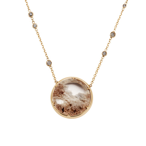 A quartz charm with a desert like scene in a bezel setting with claw prongs set in yellow gold on a yellow gold chain with grey diamonds on it.