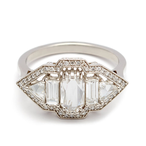 A ring with an emerald cut and shaped white diamond center stone with one baguette white diamond and one white diamond trillion on either side all encircled in white diamond pave halo set in platinum.