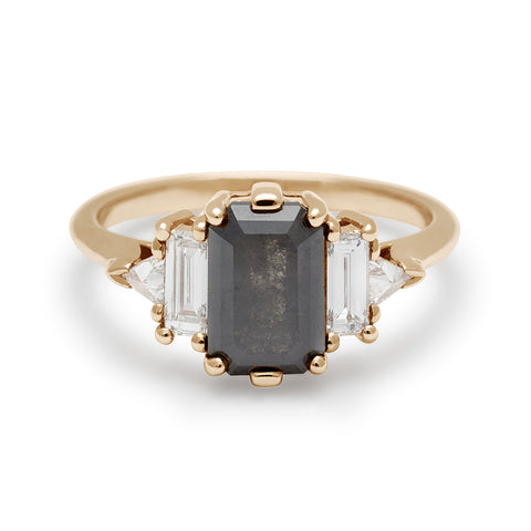 A ring with an emerald shaped black diamond center stone with one white diamond baguette and one white diamond trillion on either side set in yellow gold.