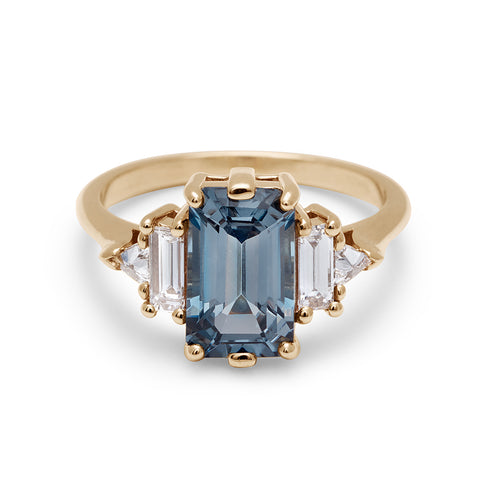 A ring with an emerald cut and shaped grey spinel center stone with one baguette white diamond and one white diamond trillion on either side set in yellow gold.