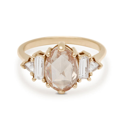 A ring with a rose cut and oval shaped champagne diamond center stone with one baguette white diamond and one white diamond trillion on either side set in yellow gold.