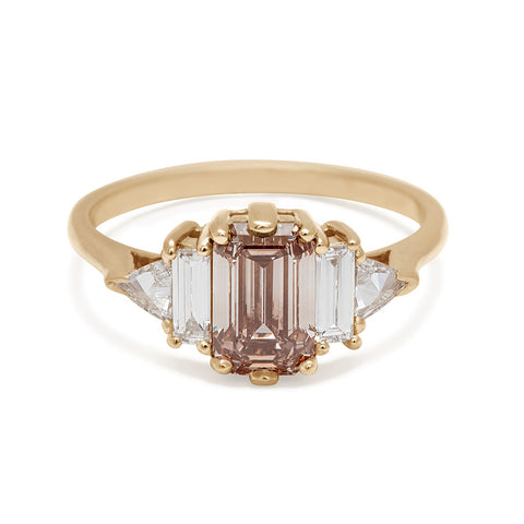 A ring with an emerald cut and shaped champagne diamond center stone with one baguette white diamond and one white diamond trillion on either side set in yellow gold.