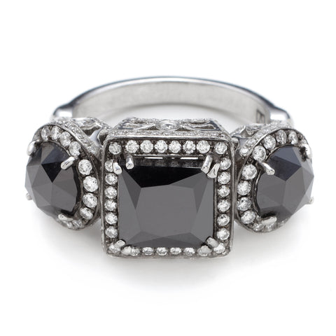 A three stone ring with an emerald cut and emerald shape black diamond center stone with two round black diamond side stones. All surrounded with white diamond pave halo set in platinum.