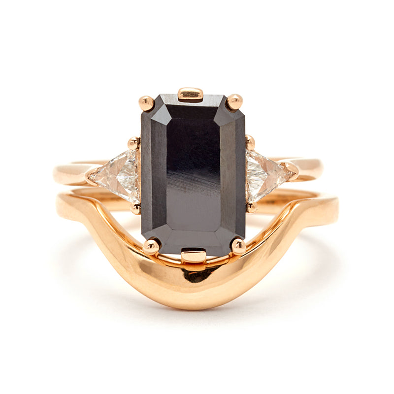 A three stone engagement ring with an 11x7mm black diamond center stone with white diamond trillions set in yellow gold and stacked on top of a yellow gold solid gold curve band.