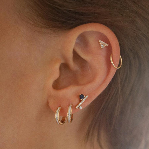 A single earring with a white trillion set above a line of white diamond pave set in yellow gold on the top of a light-complextion models ear at the base of the ear are two hoop earrings and a diamond dusted line stud., White Gold, Rose Gold, Yellow Gold 5