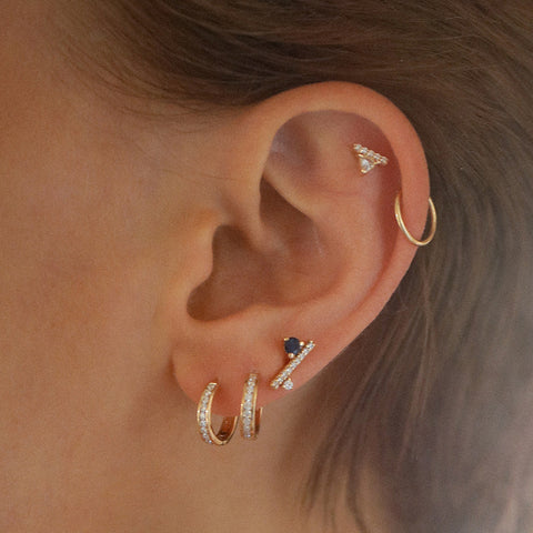 A single earring with a white trillion set above a line of white diamond pave set in yellow gold on the top of a light-complexion models ear at the base of the ear are two hoop earrings and a diamond dusted line stud., White Gold, Rose Gold, Yellow Gold 3