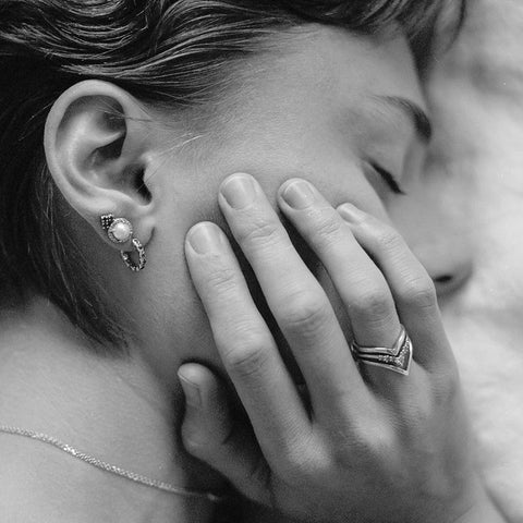 Black and white photo of a model laying down wearing two stud earrings and one huggie hoop and touching her face with her right hand. On her right ring finger she wears three nesting bands. 2